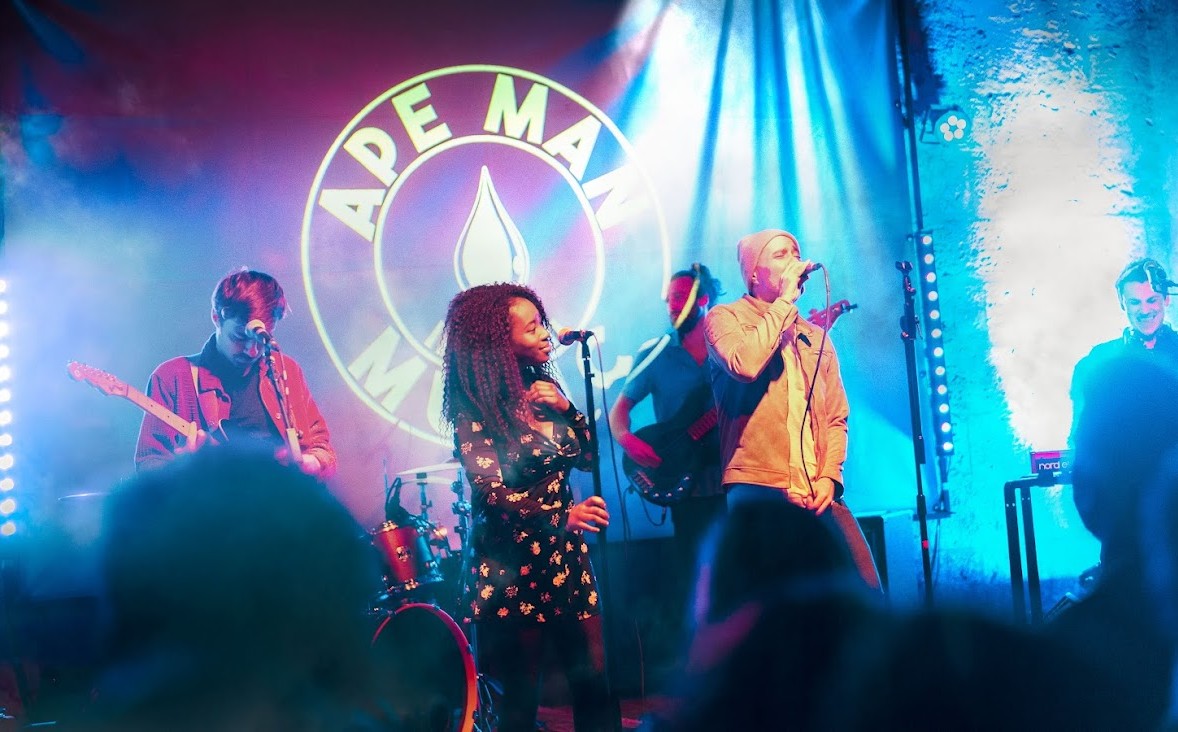 Ape Man, sextet en concert, chanteuse et chanteur au micro devant le logo du groupe